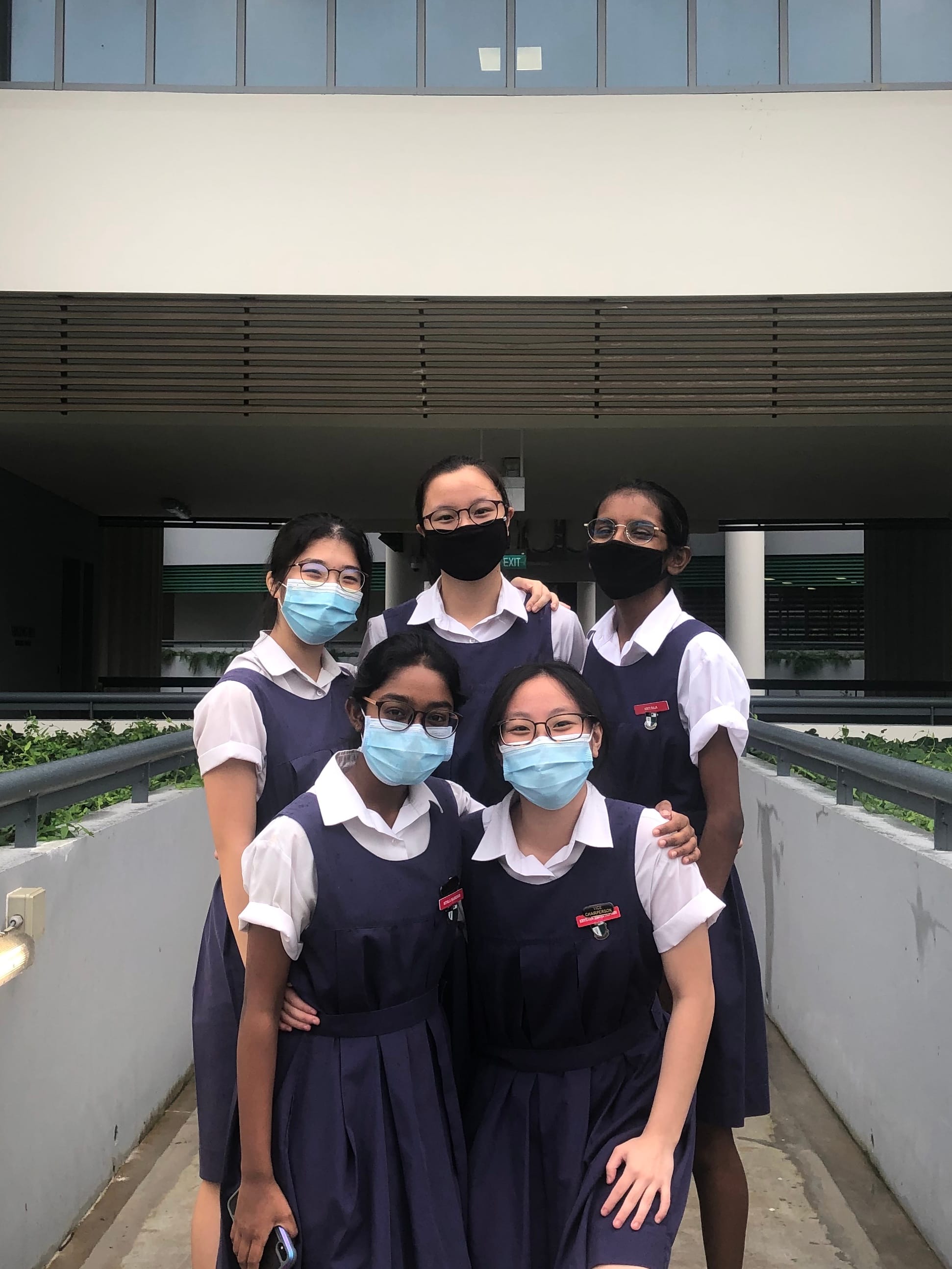 Five schoolgirls in navy uniforms and face masks pose together outside a modern building.